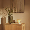 Green vase with leaf pattern, yellow table lamp, wood cabinet, and earthy tones create a warm and inviting ambiance.