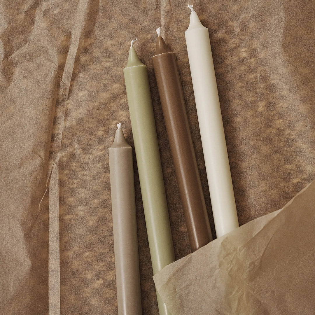 "Set of four tapered candles in earthy tones: olive green, brown, cream, and white on textured brown paper."