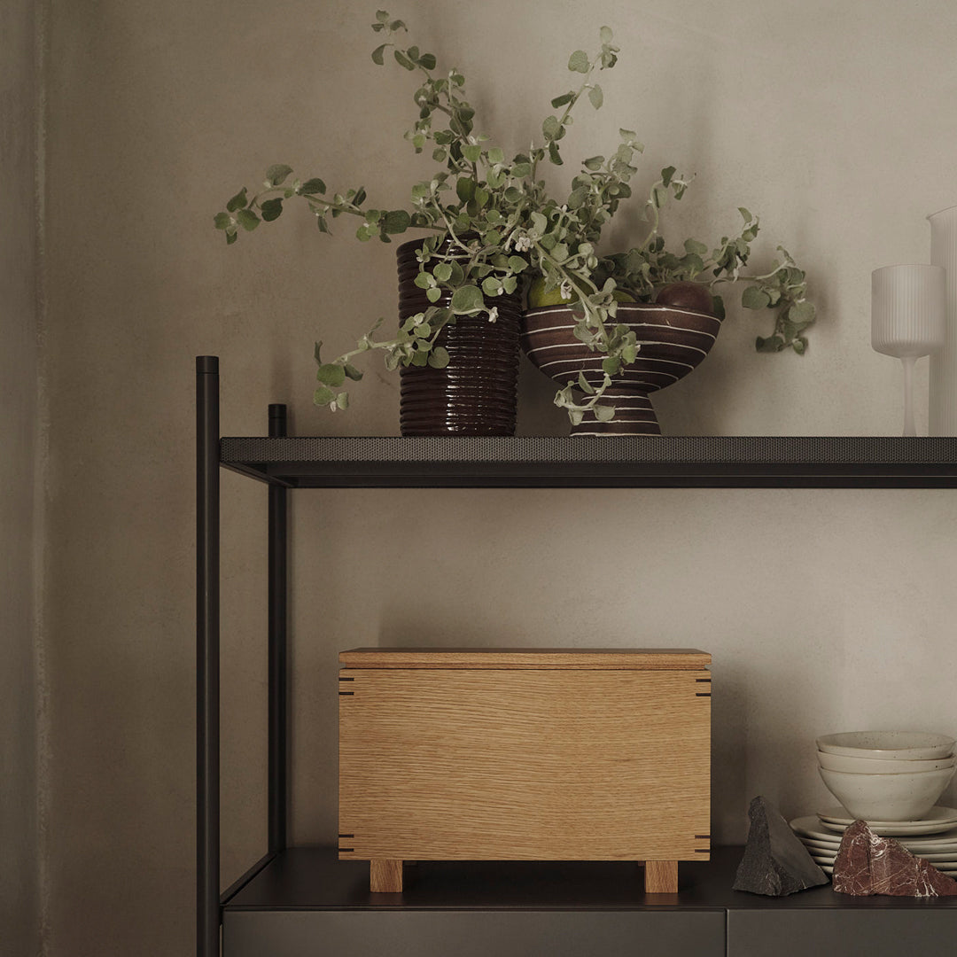 Light wood box with clean lines and natural finish, surrounded by deep brown ceramic vases and greenery accents.