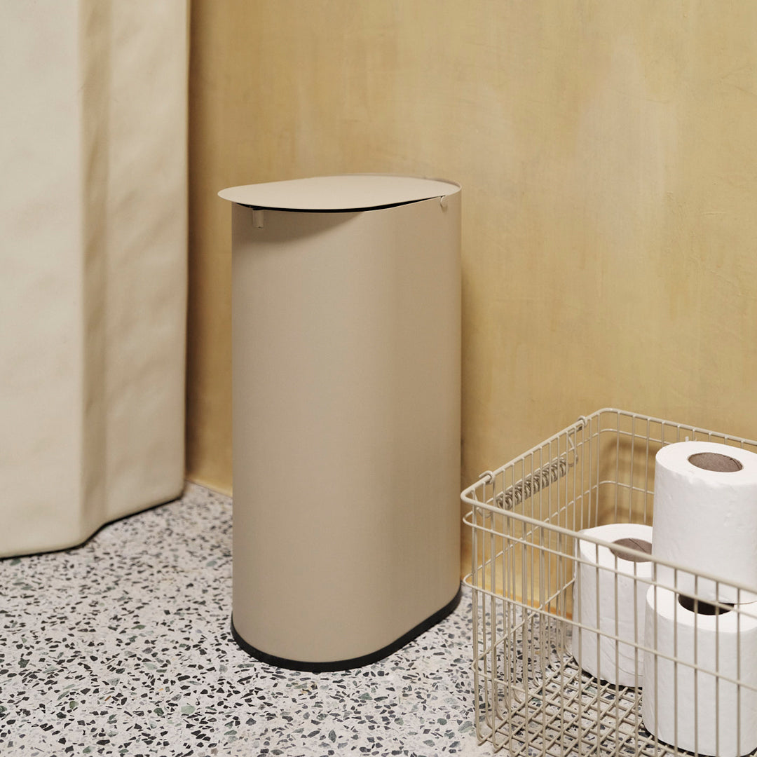 Beige cylindrical trash can with a sleek lid, set against a textured speckled floor and warm yellow wall.