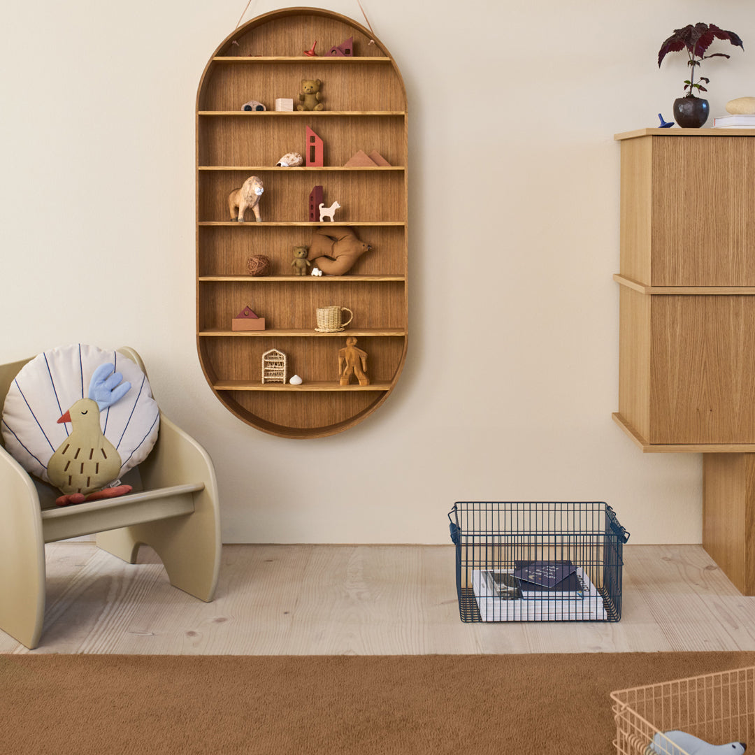Wooden bookshelf with rounded edges, neutral walls, beige armchair with colorful pillow, and a blue wire basket.
