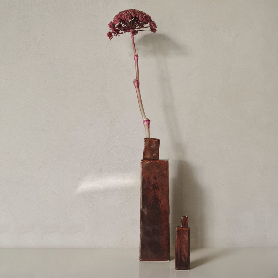 "Two geometric vases in rich brown tones with a striking pink dried flower stem, set against a neutral background."