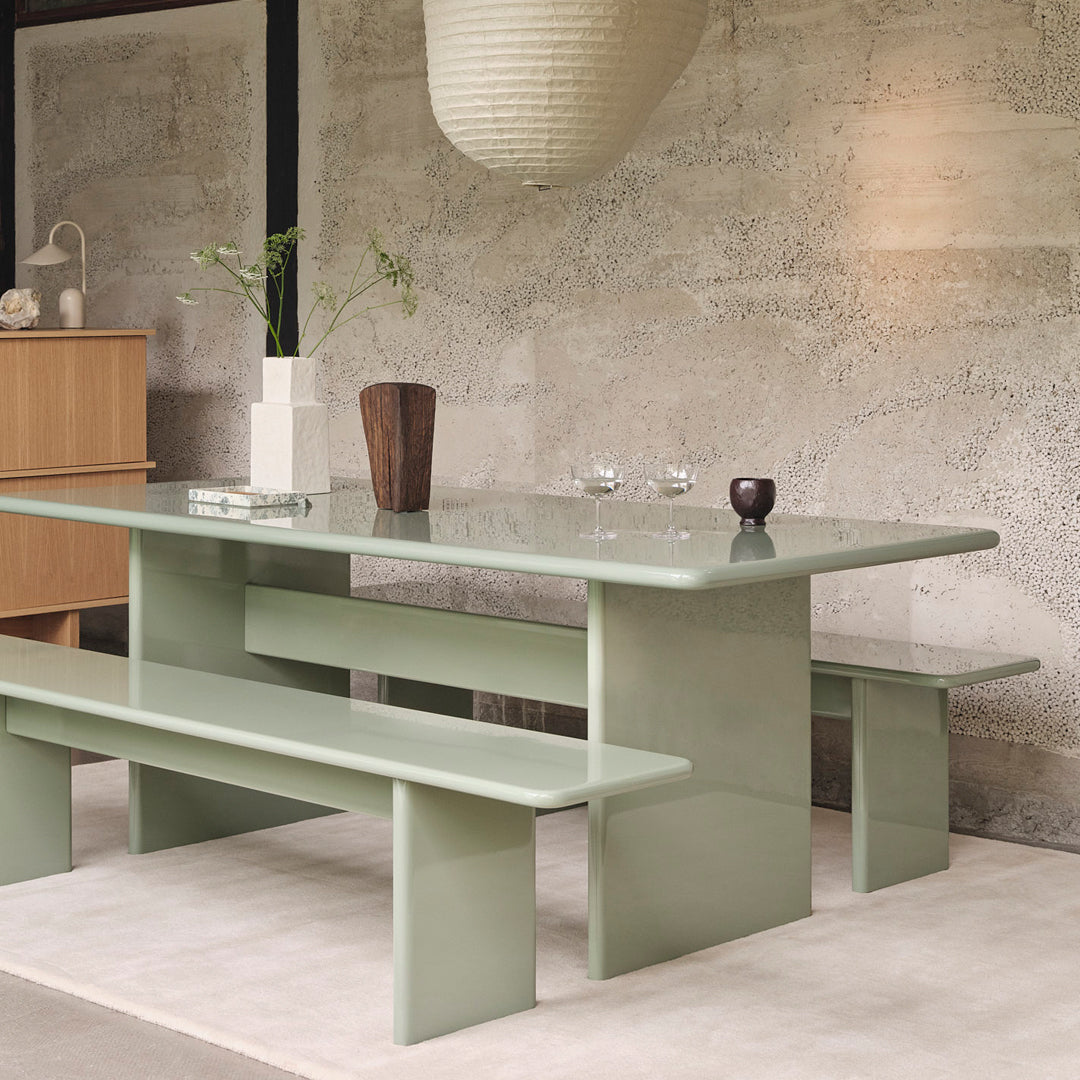 Modern green dining table and benches with a sleek, glossy surface, set against a textured stone wall backdrop.