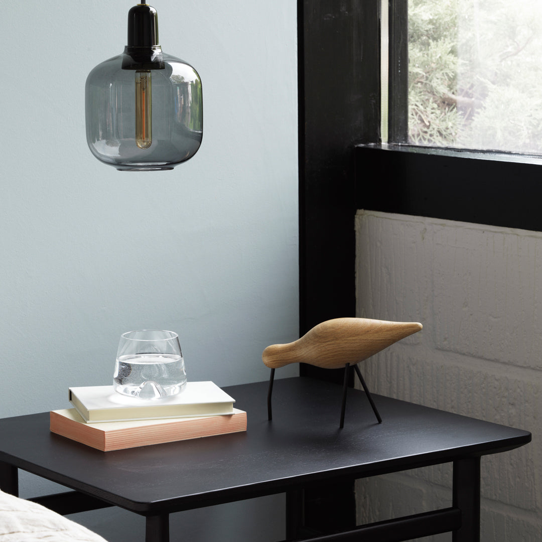 Textured black table with a smoky glass lamp, wooden bird, and stacked books in neutral tones against a soft blue wall.