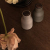 Two minimalist vases in soft gray and muted beige with wooden lids, placed on a dark wooden surface.
