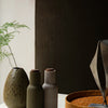 "Muted green and beige ceramic bottle vases with a textured brown tray, set against a dark grey wall."