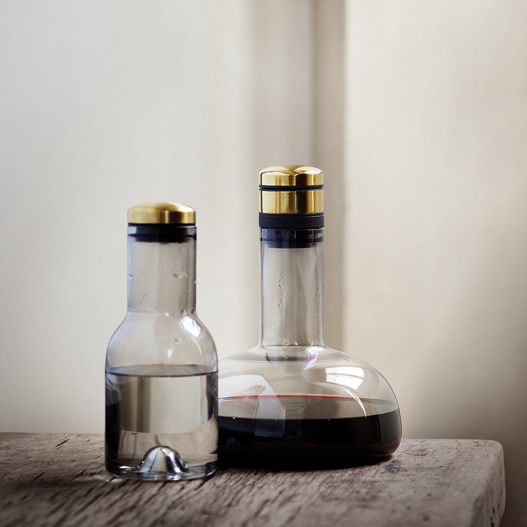 Clear glass bottles with gold and black accents, one containing water and the other red wine, on a rustic wooden surface.