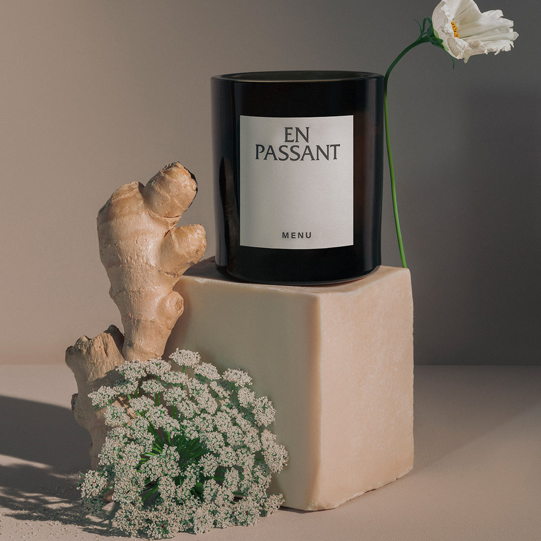 Black candle with white label on beige block, surrounded by light brown ginger, white flowers, and green foliage.