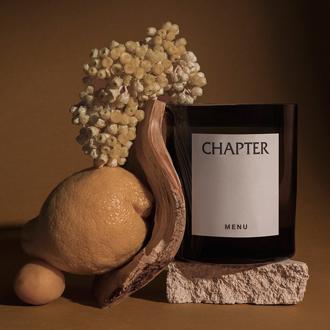 Black glass candle with a white label, surrounded by earthy browns, lemon yellow, and soft beige floral accents.