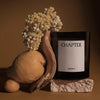 Black glass candle with a white label, surrounded by earthy browns, lemon yellow, and soft beige floral accents.