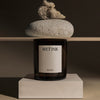 Dark amber candle jar, minimalist white label, natural gray stone, earthy tones, and soft wood elements.
