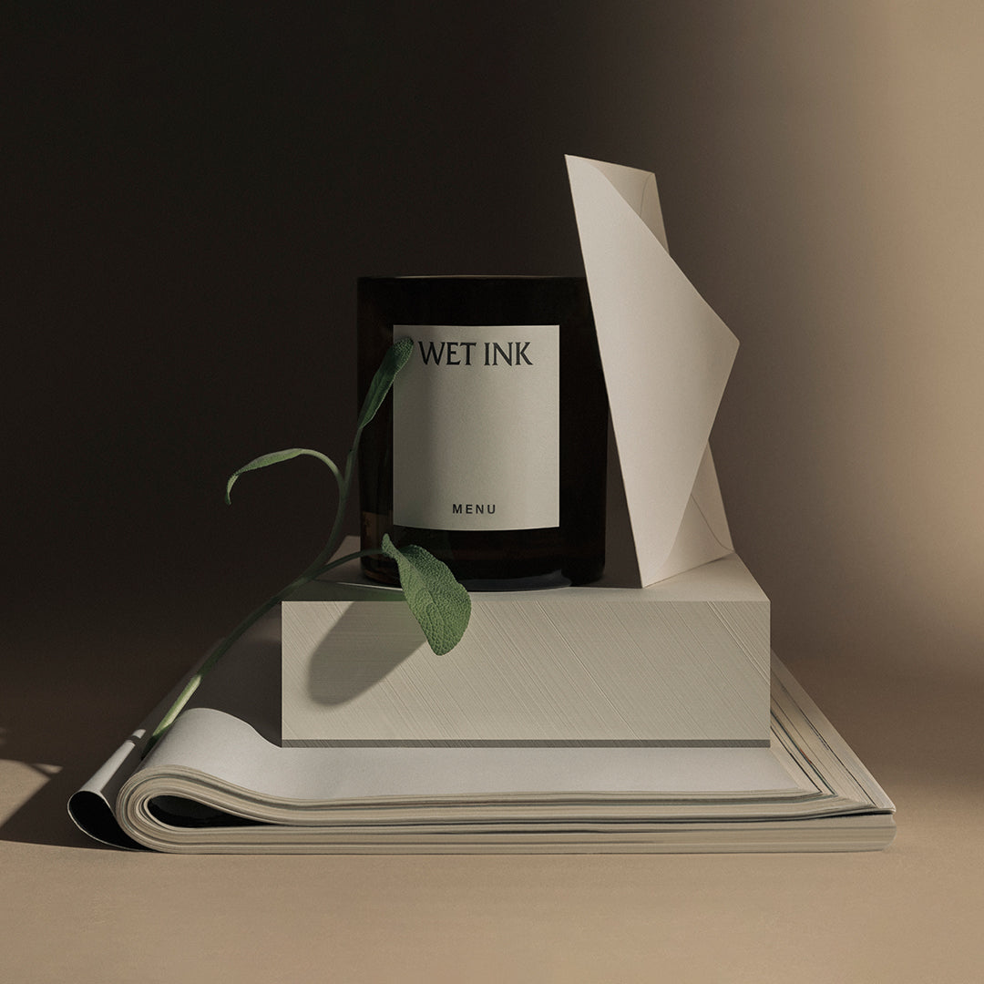 Black candle with a beige label, set against soft brown and cream background, surrounded by green foliage and textured paper.