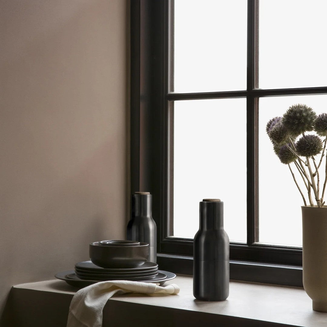Black ceramic bottles and plates on a neutral-toned table near a window, featuring subtle textures and soft, muted colors.