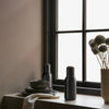 Black ceramic bottles and plates on a neutral-toned table near a window, featuring subtle textures and soft, muted colors.