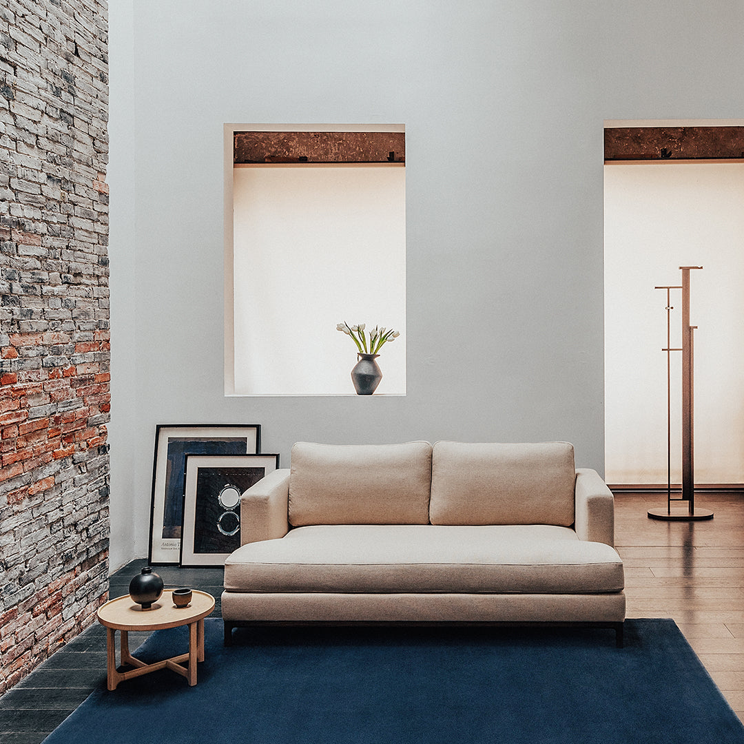 Beige sofa on a dark blue rug against a textured gray and brick wall, complemented by light-toned windows.