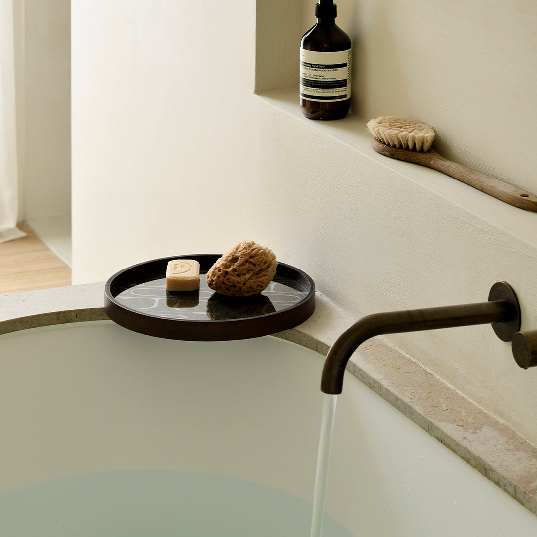 "Elegant round dark tray with natural sponges, contrasting against a light stone bath and soft beige wall."