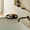 "Elegant round dark tray with natural sponges, contrasting against a light stone bath and soft beige wall."