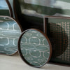 Round and rectangular trays featuring green geometric patterns and rich brown frames. Modern decor accents.