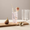 Clear glass tumbler with pink drink, gold spoon, fresh fig, and neutral wood surface; soft beige background.
