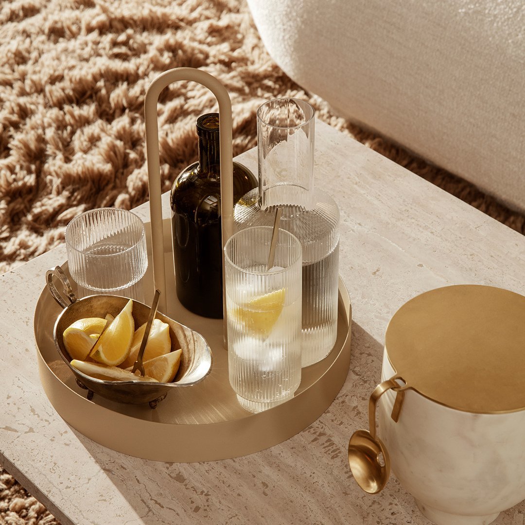Gold-toned serving tray with elegant glassware, featuring clear drinks and lemon accents on a textured beige surface.