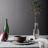 Dark purple bowl on a light gray plate, green glass bottle, and textured gray backdrop with a hint of greenery.