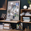 Framed corkboard with a neutral gray texture, surrounded by dark wood and soft pastel decor elements.