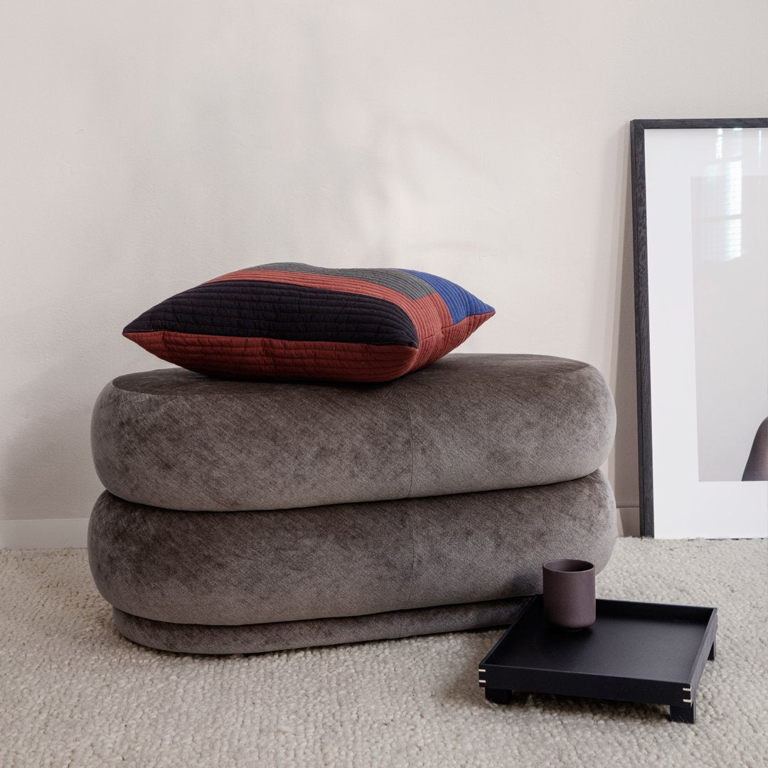 Gray plush ottoman stacked with a multi-colored striped pillow featuring red, blue, and black accents.