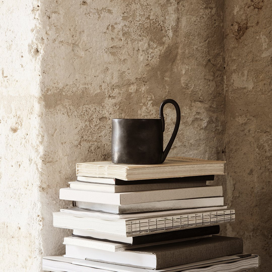 Black ceramic mug on stacked white and grey books against a textured stone wall background.