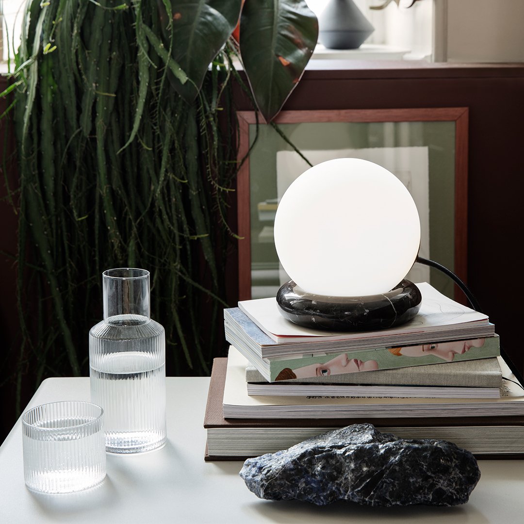 Minimalist lamp with a white globe on a dark base, surrounded by layered pastel books and clear glassware against a green backdrop.