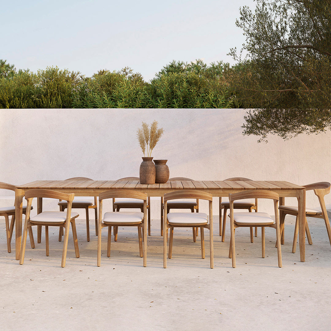 Natural wood dining table with light beige cushions, set against a soft white wall and green foliage backdrop.