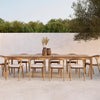 Natural wood dining table with light beige cushions, set against a soft white wall and green foliage backdrop.