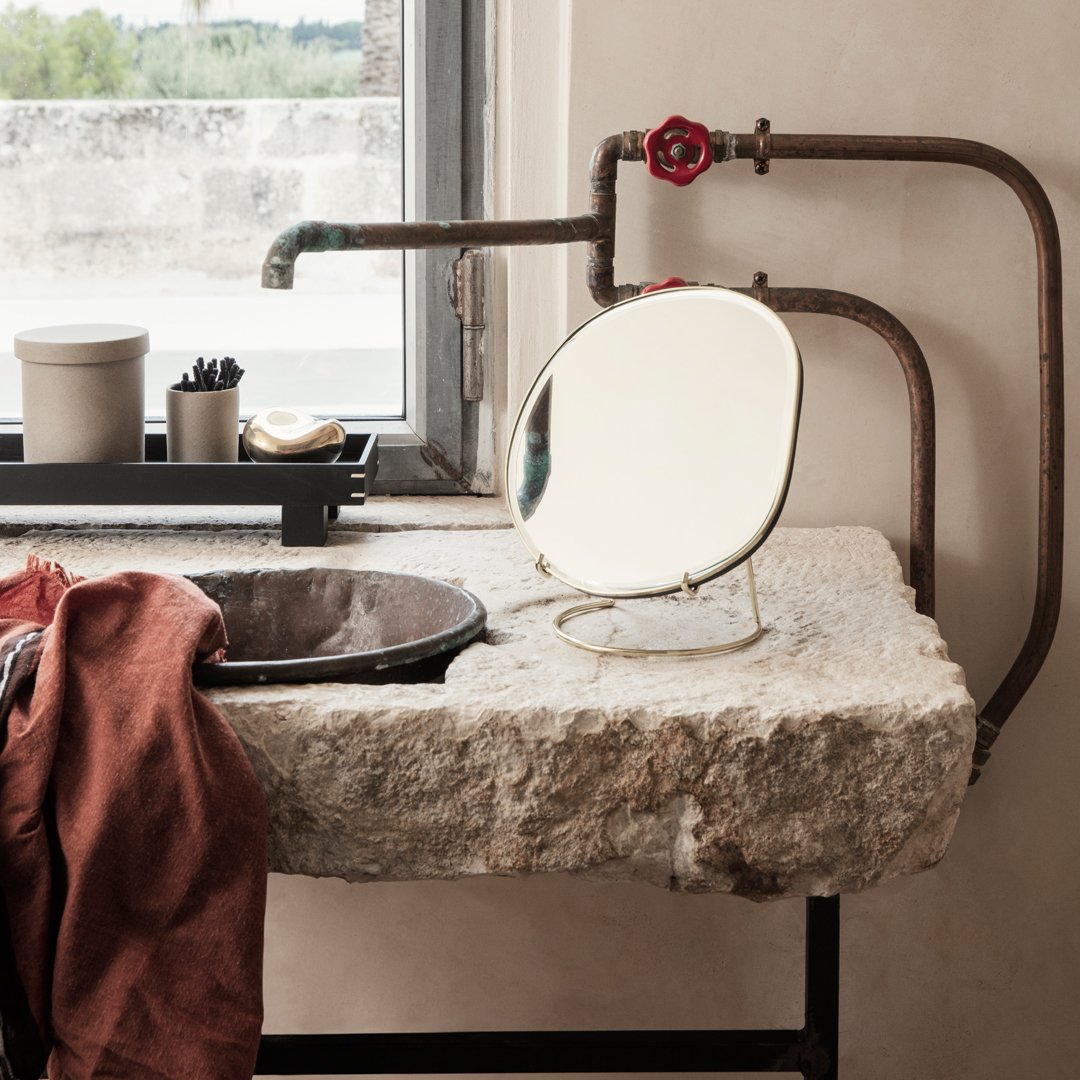 Round gold mirror on a rustic stone sink with a brown cloth and industrial metal accents in a natural light setting.