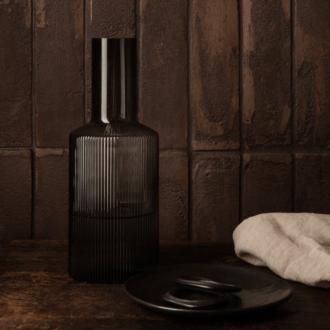 Black ribbed glass vase with a sleek cylindrical neck, set against a rustic brown backdrop and paired with dark dishware.