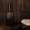 Black ribbed glass vase with a sleek cylindrical neck, set against a rustic brown backdrop and paired with dark dishware.