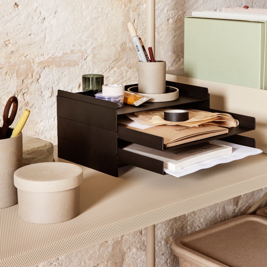 Black wooden organizer with layered shelves, set against a textured beige wall, featuring earthy brown and green accessories.