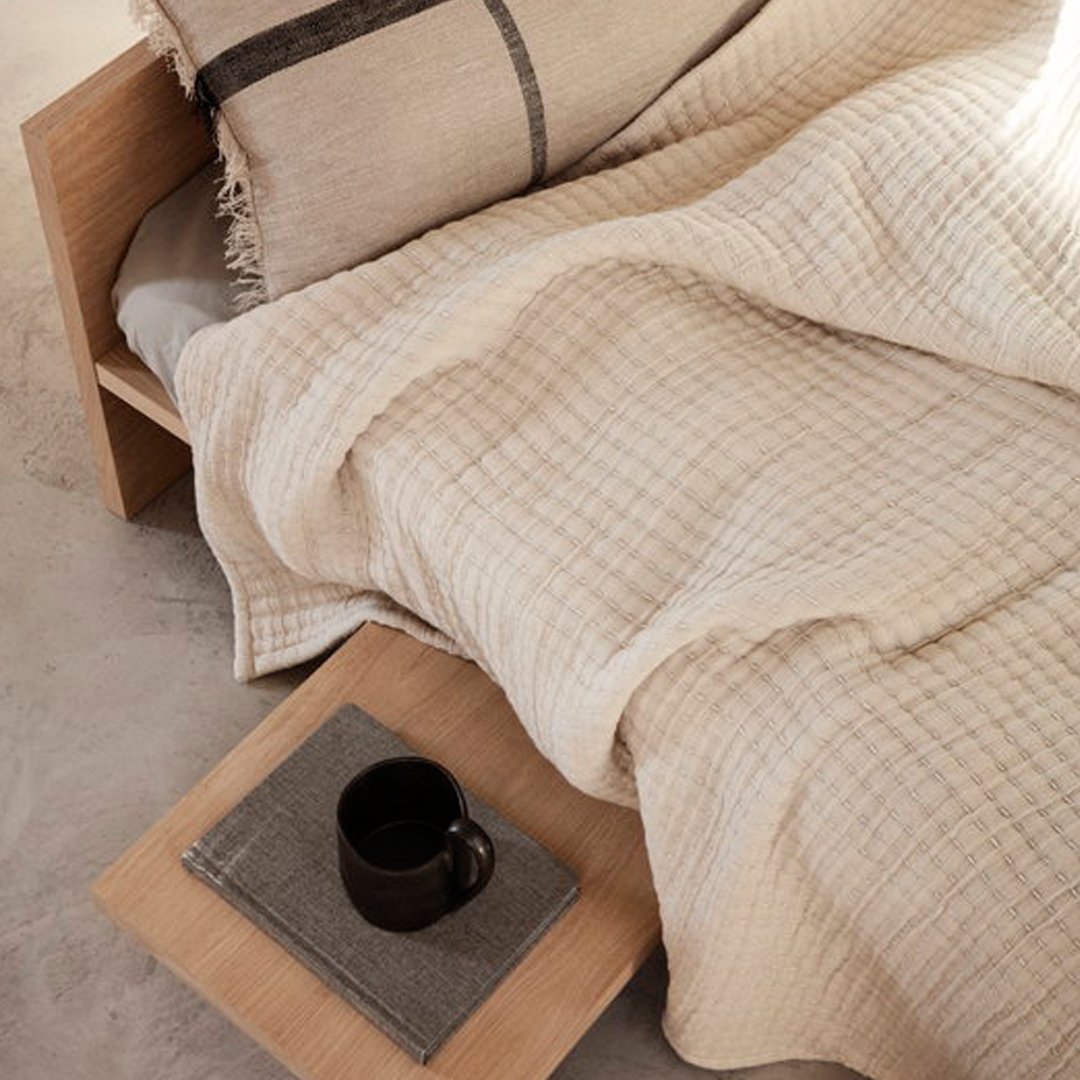 Beige textured blanket with a subtle grid pattern, paired with a neutral pillow and wooden bedside tray.
