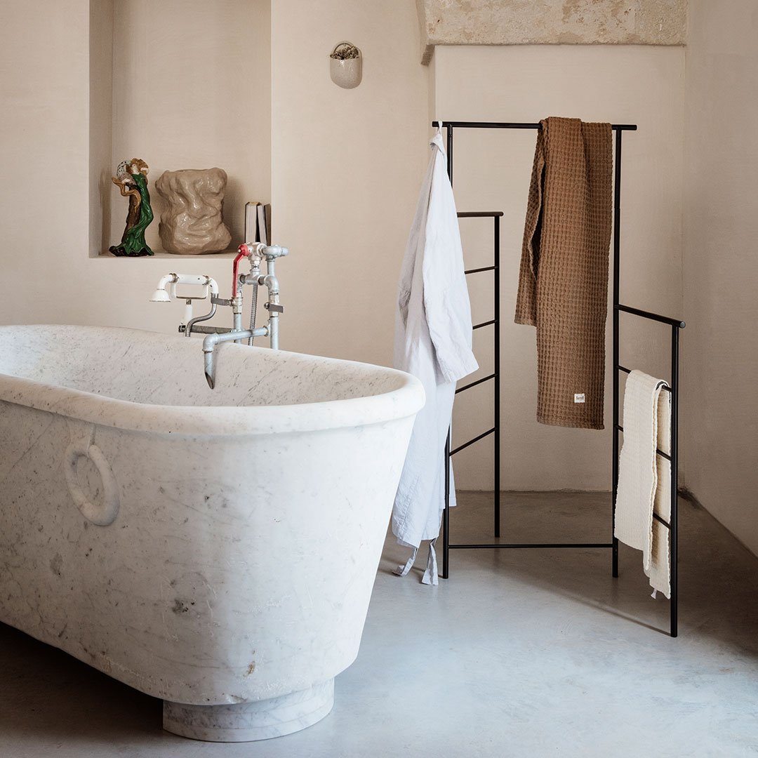 Elegant marble freestanding bathtub with a minimalist black rack displaying beige and white towels in a neutral-toned bathroom.