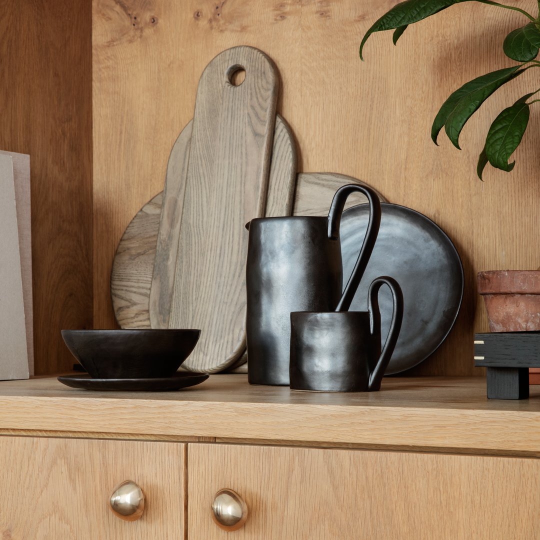 Black ceramic dishes and polished metal accents against natural wood textures. Elegant and modern tableware display.