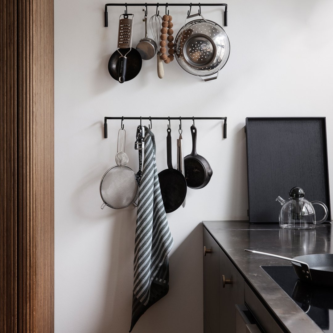 Black kitchen hangers with silver utensils and a gray striped towel against a minimalist white wall.
