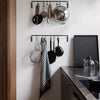 Black kitchen hangers with silver utensils and a gray striped towel against a minimalist white wall.