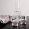 Clear ribbed glass water carafe and tumblers with fresh herbs and berries, set on a dark wooden table with a textured towel.