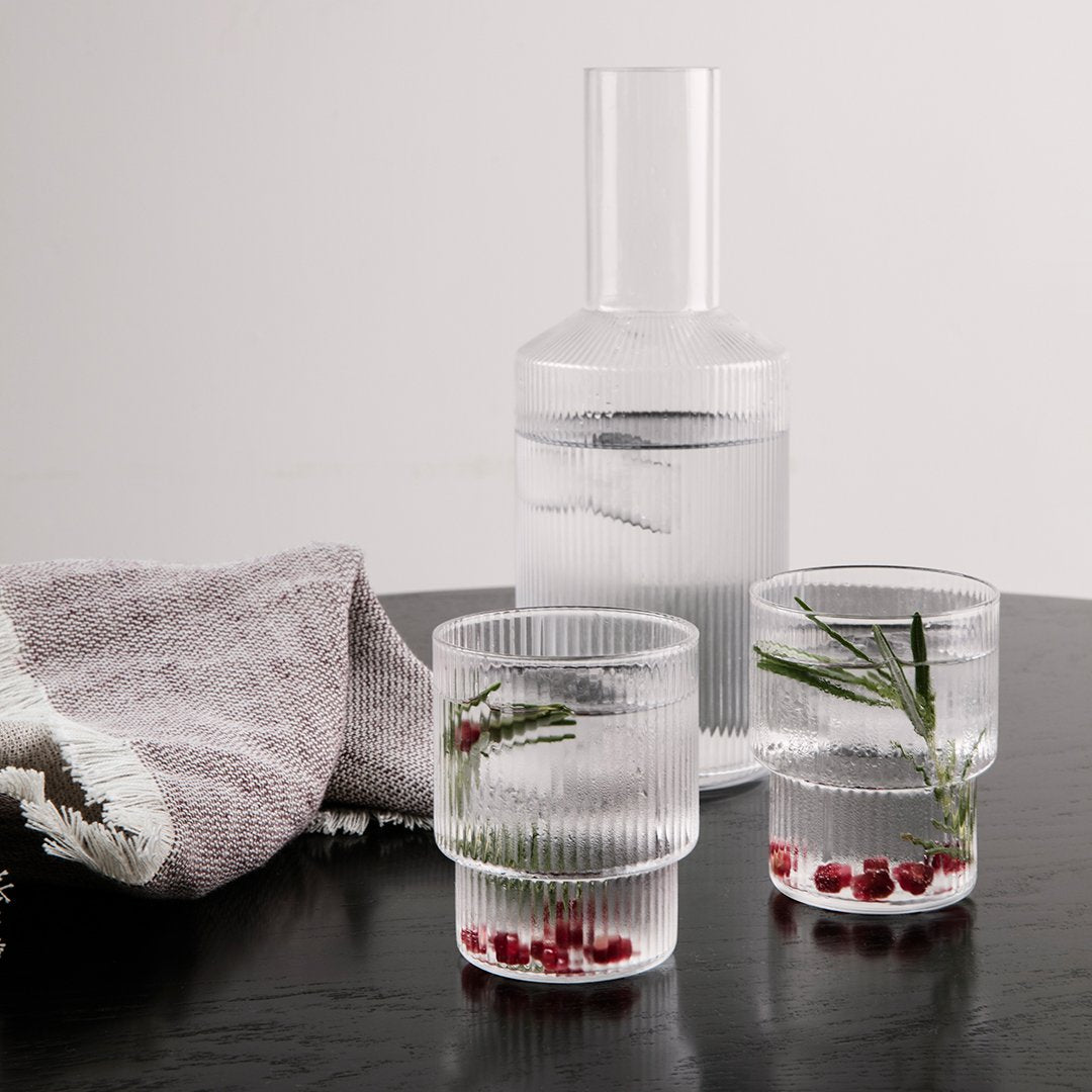 "Clear ribbed glass pitcher and glasses with green herbs and red accents on a wooden table, complemented by a textured cloth."