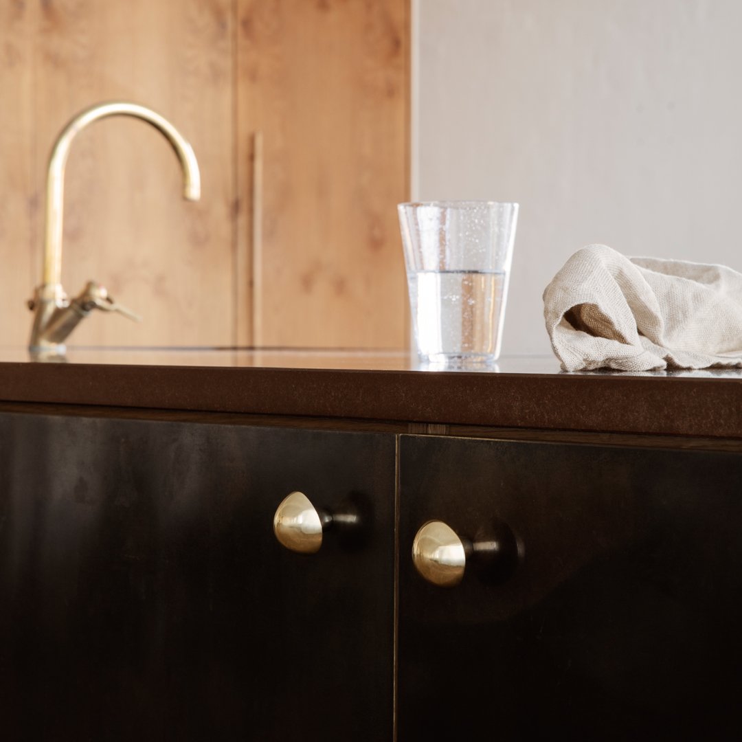 Dark wooden cabinet with brass knobs, topped with a light-colored cloth and a clear glass of water, against natural wood backdrop.