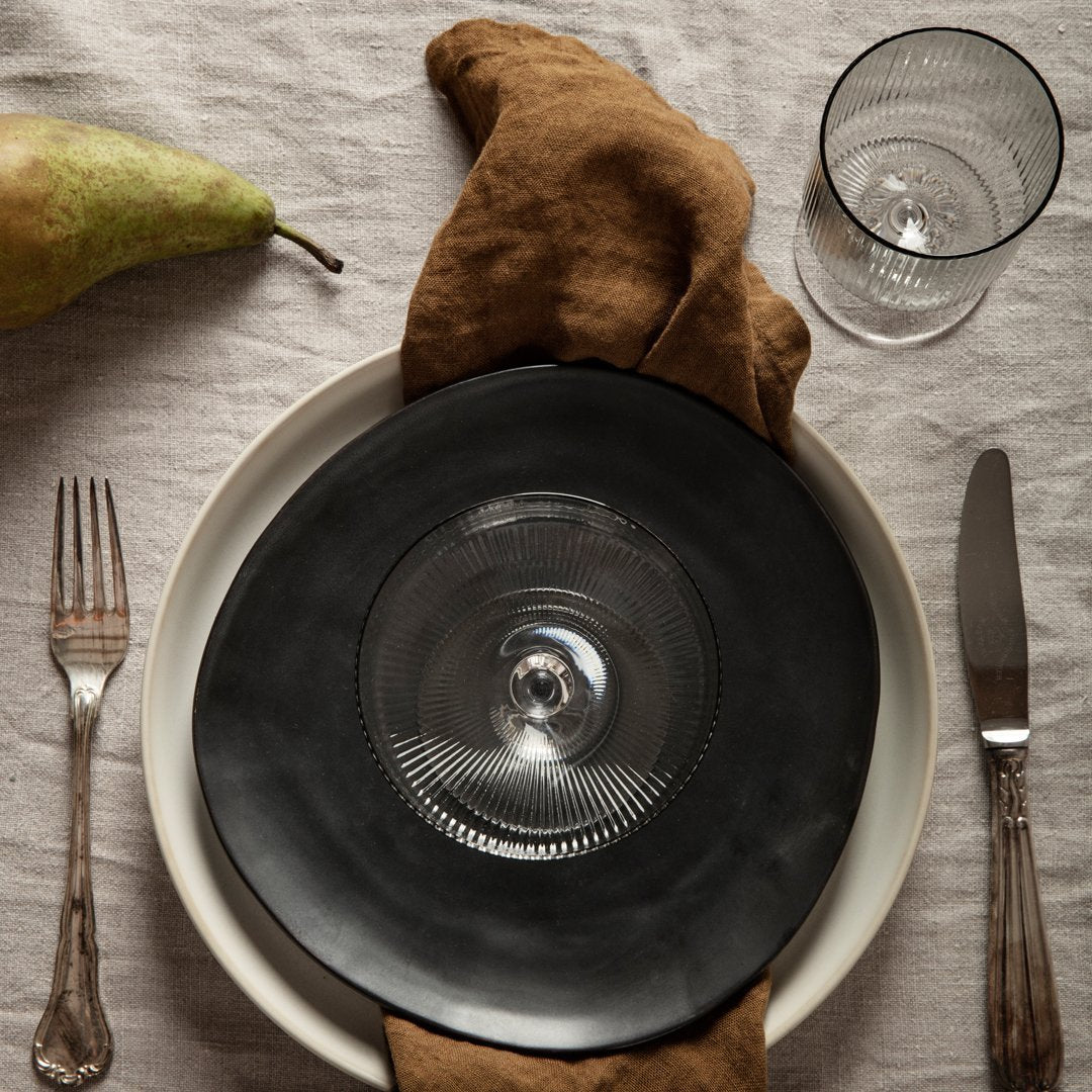 "Elegant black plate with intricate pattern, paired with cream tableware and brown linen napkin, set on a textured neutral tablecloth."