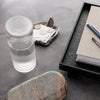 Clear ribbed glass tumbler on a textured gray surface with rustic stone and sleek black tray elements.