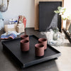 Rustic black tray with four maroon cups on a dark countertop, featuring natural textures and a minimalist aesthetic.