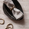 Linen napkin wrapped in a gold ring on a textured black ceramic plate; neutral tones and elegant shapes.