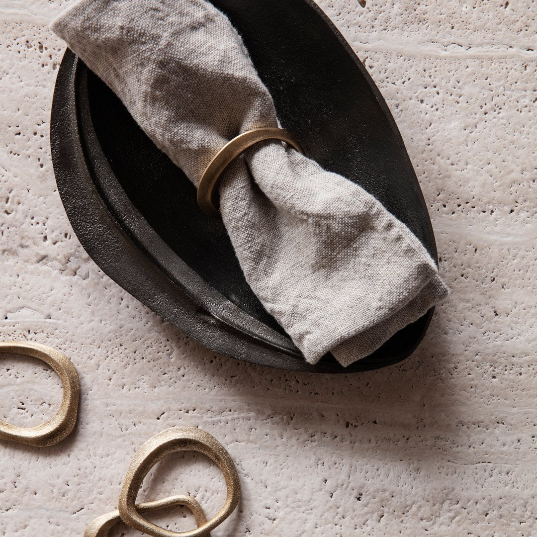 "Neutral gray linen napkin wrapped with a gold ring, set on a matte black ceramic plate, featuring organic shapes."
