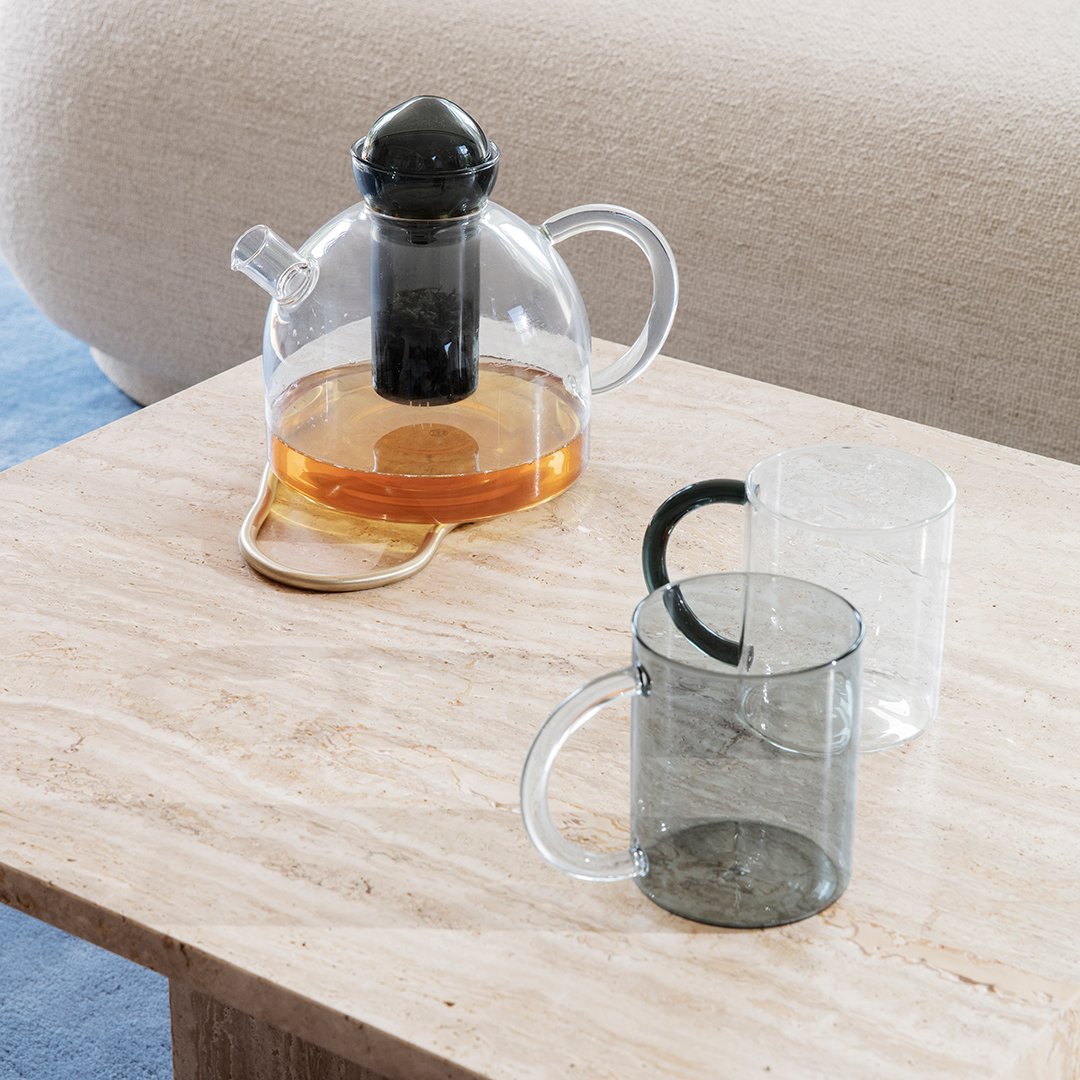 Clear glass teapot with black lid and smokey gray mugs on a light marble table. Warm tea color contrasts with cool tones.