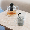 Clear glass teapot with black lid and smokey gray mugs on a light marble table. Warm tea color contrasts with cool tones.